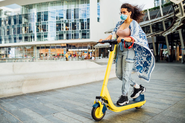 Jovem mulher de raça mista em máscara facial montando scooter elétrico na cidade
  - Foto, Imagem