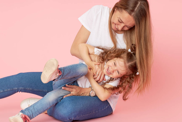 Family values. Mom and daughter in white t-shirts and jeans play and hug on a pink background. Caring for loved ones. Happy motherhood - Photo, image
