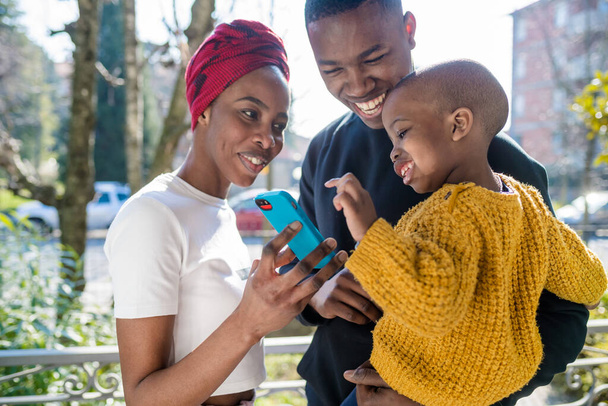černá šťastná rodina venku pomocí chytrého telefonu s úsměvem - technologie, mládež kultura, rodinný koncept - Fotografie, Obrázek