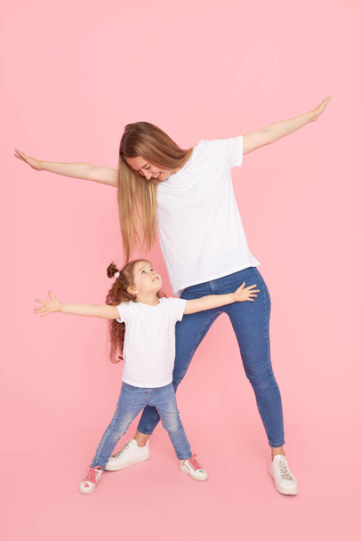 Family values. Mom and daughter in white t-shirts and jeans play and hug on a pink background. Caring for loved ones. Happy motherhood - Foto, immagini
