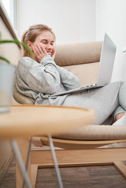 Happy woman in gray home suit with laptop in her hands sitting in chair - Фото, зображення