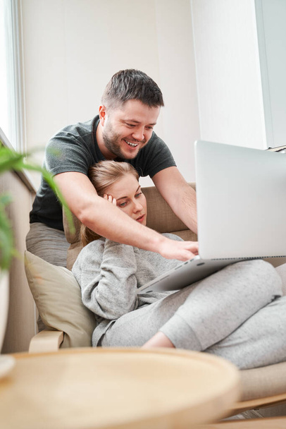 Young blonde sitting on beige armchair with laptop and man standing near her in apartment - Foto, imagen