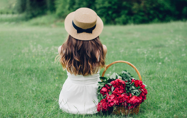 Beyaz elbiseli bir kız sırtı yeşil çimlerin üzerinde oturuyor sepetin yanındaki kırmızı şakayıklı. - Fotoğraf, Görsel