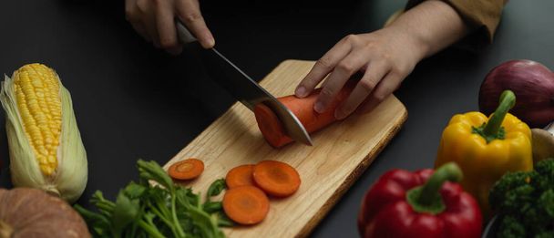 Tiro cortado de cozinheiro fêmea cortando cenoura fresca no bloco de corte na cozinha moderna escura
  - Foto, Imagem