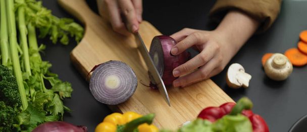 Przycięty strzał kucharki cięcia świeżej czerwonej cebuli na bloku siekania w ciemnej nowoczesnej kuchni  - Zdjęcie, obraz