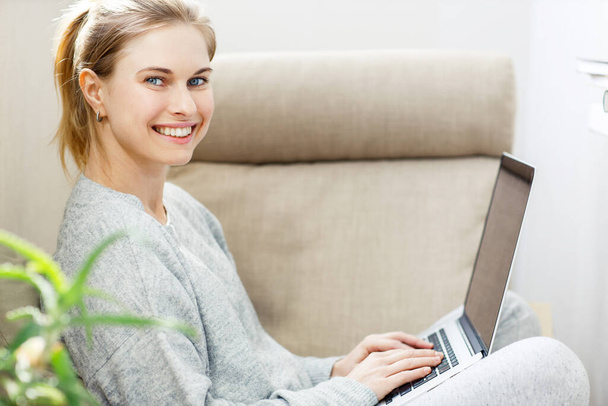Happy blonde woman in gray home suit with laptop in her hands sitting in chair - Foto, Imagem