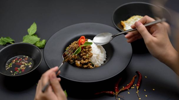 Καλλιεργημένο πλάνο του θηλυκό τρώει ταϊλανδέζικο παραδοσιακό φαγητό, Ανακατέψτε τηγανητό κιμά χοιρινό με βασιλικό (Pad ka prao) σε μαύρο τραπέζι στο ταϊλανδέζικο εστιατόριο - Φωτογραφία, εικόνα