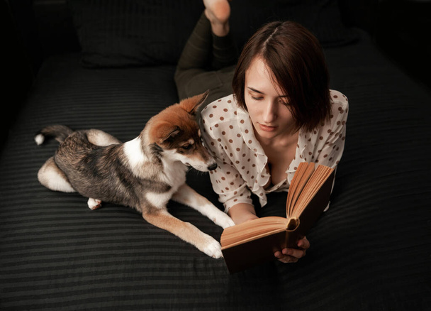 Girl with a dog lies in a black bed and reads a book - Foto, imagen