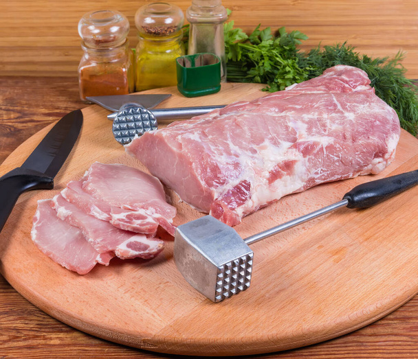 Partly sliced uncooked pork loin, kitchen knife, different meat hammers on the wooden cutting board, spices and greens on the rustic table - Photo, Image