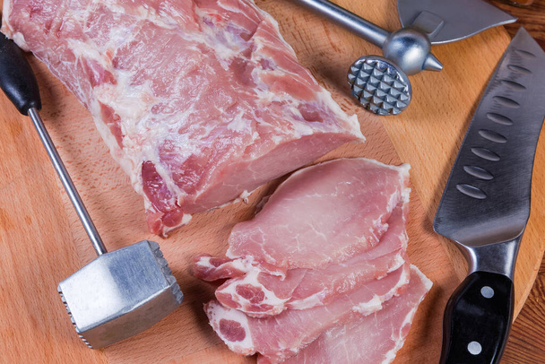 Filet de porc partiellement tranché non cuit parmi les différents attendrisseurs de viande et couteau de cuisine sur la planche à découper en bois, vue sur le dessus des fragments - Photo, image