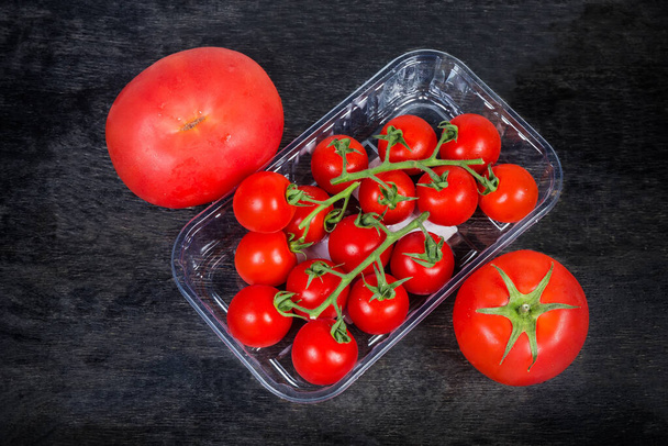 Açık tek kullanımlık plastik ambalajlı kırmızı kiraz domatesleri ve siyah yüzeyli sıradan domatesler. - Fotoğraf, Görsel