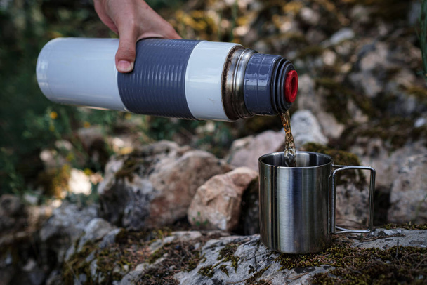 In a camping mug, a female hand pours tea from a thermos in nature. The idea and concept of a break, rest and travel - 写真・画像