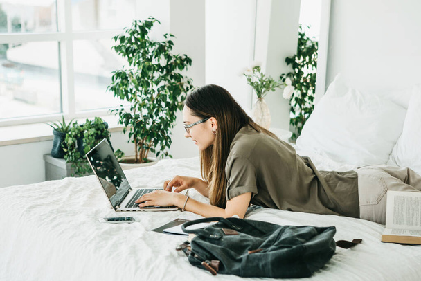 Girl student or freelancer is lying in bed and uses a laptop. Or she works from home. She is typing a text or message or is she surfing the Internet online. - Foto, Imagem