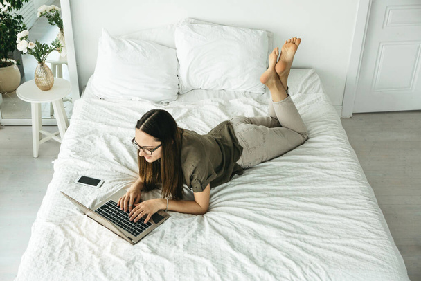 Chica positiva estudiante se encuentra en la cama y utiliza un ordenador portátil. Ella está enviando mensajes de texto o navegar en línea en Internet. - Foto, Imagen