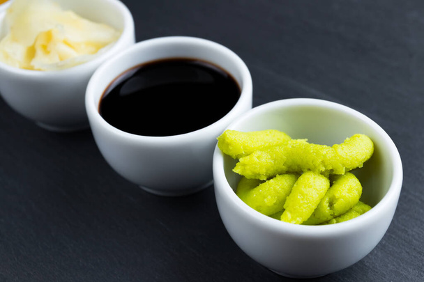 Molho de soja, gengibre, wasabi japonês rábano em pequenas tigelas brancas em fundo placa de ardósia cozinha preta. Típico asiático cozinha especiarias
. - Foto, Imagem