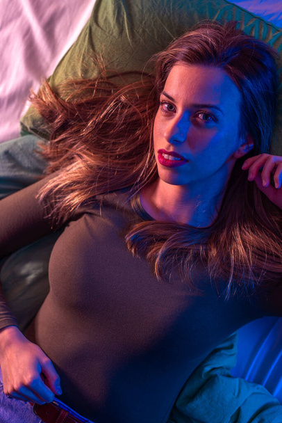 Portrait of a young woman on a bed from above with crossed colors from neon lights - Photo, Image