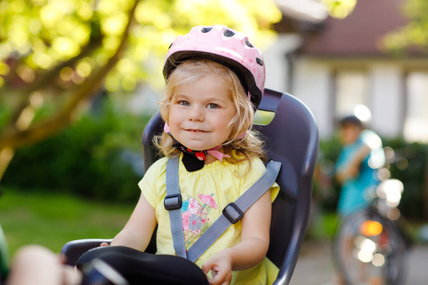 Portret van kleuter meisje met veiligheidshelm op het hoofd zittend in fietsstoeltje van ouders. Jongen op de fiets op de achtergrond. Veilig en kinderbescherming concept. Familie en weekendje weg. - Foto, afbeelding