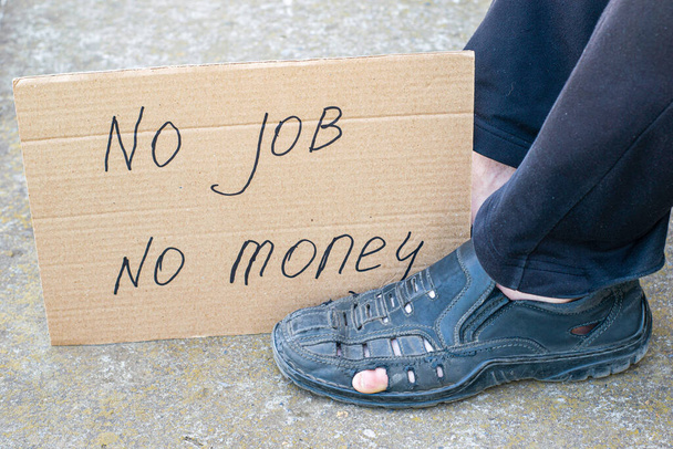 Werkloosheid concept man in oude gescheurde schoenen zit op de grond aan zijn voeten kartonnen bord zegt geen baan geen geld - Foto, afbeelding
