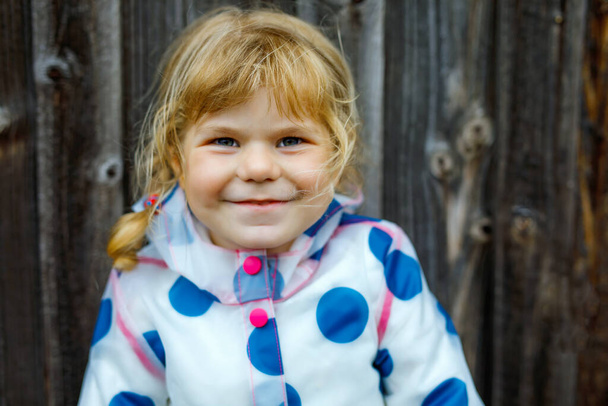 Портрет счастливой улыбающейся маленькой девочки в дождевой куртке в дождливый облачный день. Симпатичный здоровый ребенок в красочной одежде
 - Фото, изображение