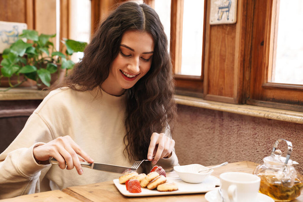 Bild einer glücklichen schönen jungen Frau, die lächelt und Pfannkuchen isst, während sie im gemütlichen Café drinnen sitzt - Foto, Bild
