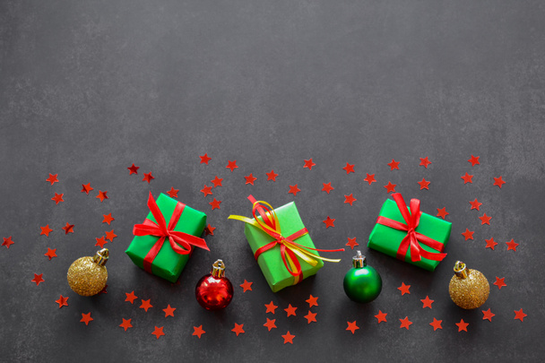 Cadeaus in groen papier met rode en gele linten, goud, rode en groene kerstballen en confetti in de vorm van rode sterren liggen op een rij. Zwarte achtergrond, kopieer ruimte. Groot verkoopconcept / - Foto, afbeelding
