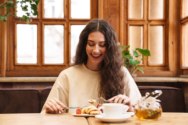 Imagen de la hermosa mujer joven feliz sonriendo y comiendo panqueques mientras está sentado en el acogedor café en el interior
 - Foto, Imagen