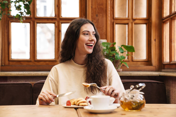 Image of happy beautiful young woman smiling and eating pancakes while sitting in cozy cafe indoors - Photo, Image