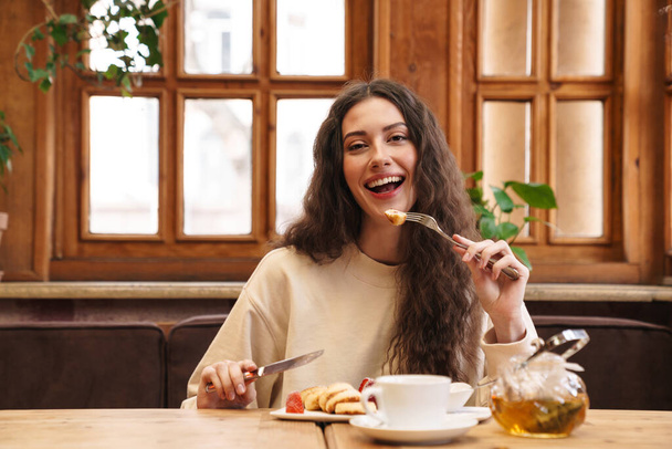 Image of happy beautiful young woman smiling and eating pancakes while sitting in cozy cafe indoors - Fotografie, Obrázek