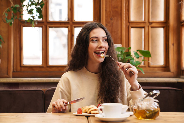 Зображення щасливої красивої молодої жінки, яка посміхається і їсть млинці, сидячи в затишному кафе в приміщенні
 - Фото, зображення