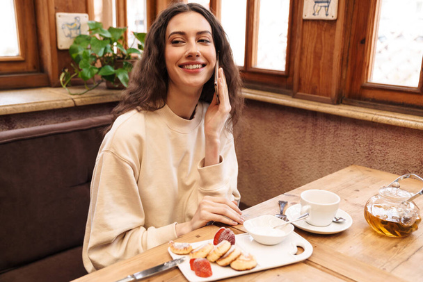 Image of joyful beautiful woman talking on cellphone while having breakfast in cozy cafe indoors - Foto, imagen