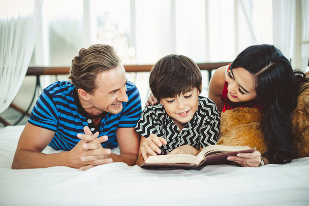 母、父と息子、かわいい少年読書本、白いベッドで宿題を教える、幸せな家族とライフスタイルの概念 - 写真・画像