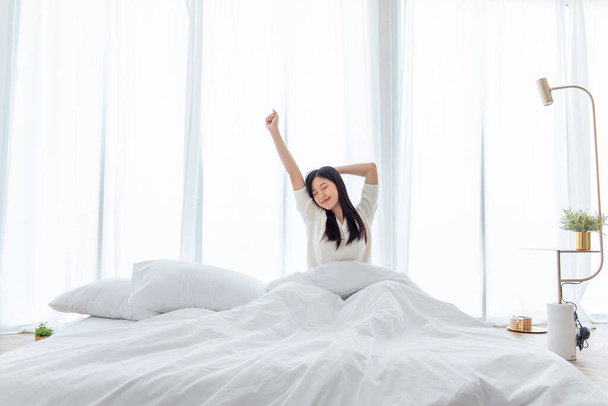 Mujer asiática despierta por la mañana, sentada en la cama blanca y estirándose, sintiéndose feliz y fresca - Foto, imagen
