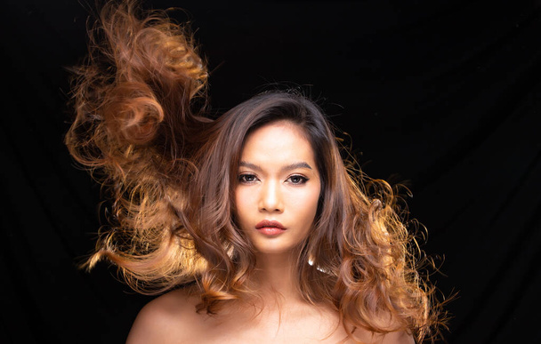 Retrato Head shot de la piel bronceada de los años 20 joven mujer asiática pelo rubio y soplar pelo revoloteando en el aire con el ventilador, mira la cámara sobre fondo de cortina de drapeado negro
 - Foto, Imagen