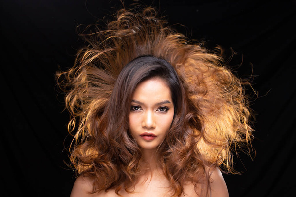 20 'li yaşlarda bronz tenli genç Asyalı kadın saçı ve dalgalı saçlarını hava üfleyicisiyle havaya savururken çekilmiş bir portre resmi. Siyah perde arkasındaki kameraya bakın. - Fotoğraf, Görsel