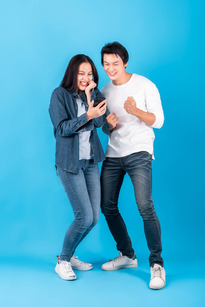 Dwóch młodych Azjatów para mężczyzna i kobieta za pomocą telefonu komórkowego ze szczęśliwą i wesołą niespodzianką na niebieskim tle, studio shot full length - Zdjęcie, obraz