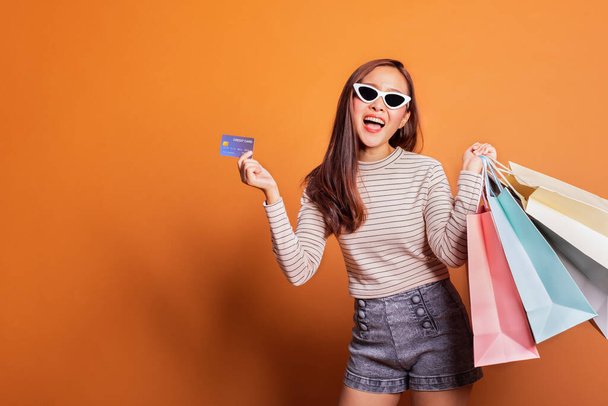 Young beautiful fashionable Asian woman holding shopping bags phone and credit card over orange background studio shot - Foto, Imagen