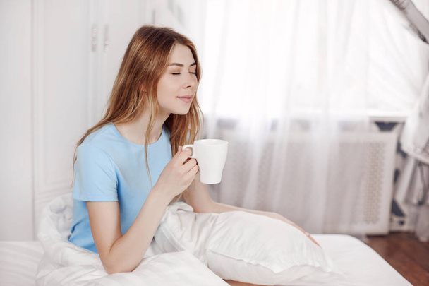 Junges schönes Mädchen trinkt Morgenkaffee sitzend im Bett. Das Mädchen genießt den Morgen, hält eine Tasse mit einem heißen Getränk in der Hand - Foto, Bild