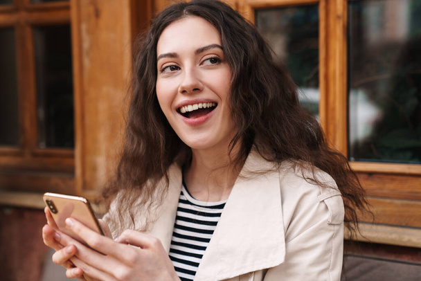 Image of happy brunette woman using mobile phone and smiling while sitting in street cafe outdoors - Фото, изображение
