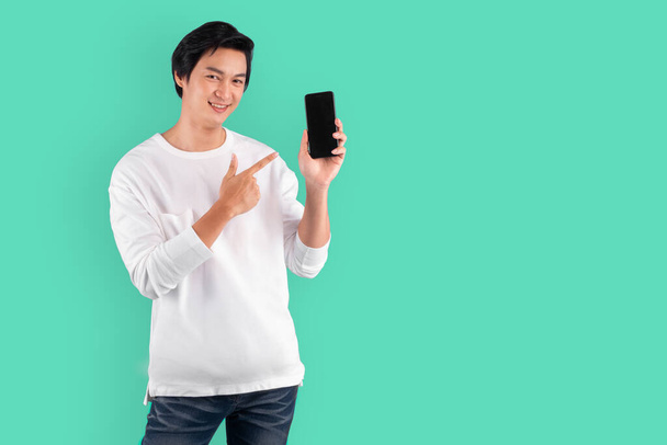 Giovane uomo asiatico che punta al telefono cellulare con felice e allegro su sfondo verde con spazio copia - Foto, immagini