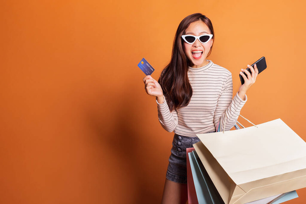 Joven hermosa mujer asiática de moda sosteniendo bolsas de compras teléfono y tarjeta de crédito sobre naranja fondo estudio disparo - Foto, Imagen