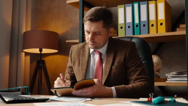 A man in a suit makes notes in a notebook. Businessman in office sitting at desk writes. - Metraje, vídeo