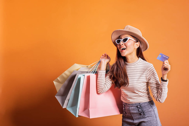 Young beautiful fashionable Asian woman holding shopping bags phone and credit card over orange background studio shot - Photo, Image
