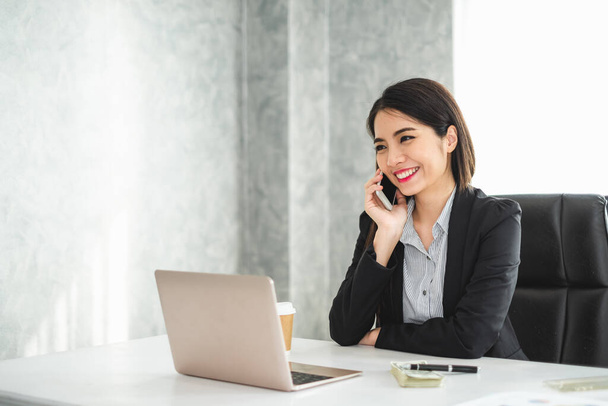 Молодая красивая азиатская предпринимательница разговаривает по телефону и работает за столом в современном офисе
 - Фото, изображение