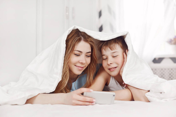 A beautiful young girl and her cheerful younger brother look at photos on their phone while lying on the bed with a smile. Brother and sister watch videos on their phone. Friendship, care, support. Space for text - Foto, Imagem