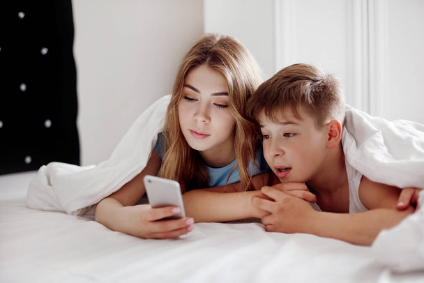 A young girl shows her younger brother a video on her phone while lying on a bed. Brother and sister watch videos on their phone. Friendship, care, support. Space for text - Photo, Image