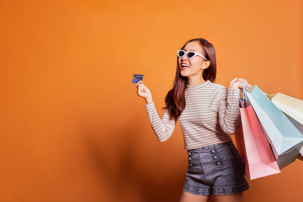 Young beautiful fashionable Asian woman holding shopping bags phone and credit card over orange background studio shot - 写真・画像
