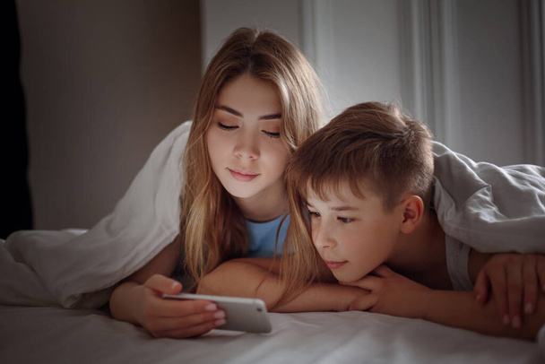 A young girl shows her little brother a video on her phone while lying on a bed at night. Brother and sister are watching a video on their phone. Friendship, care, support. Space for text - 写真・画像