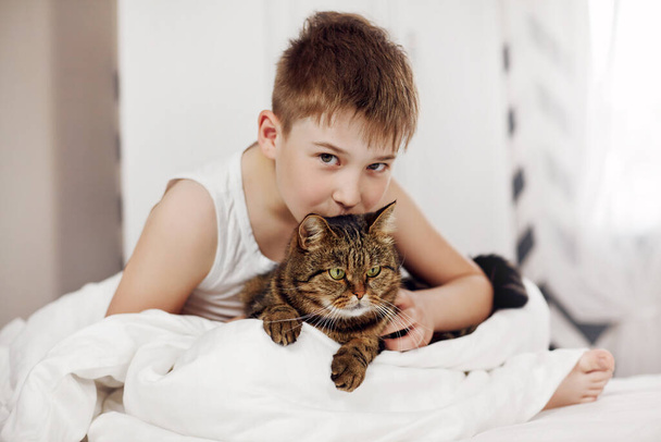 Un lindo adolescente está sentado en la cama y sosteniendo un gran gato en sus brazos. Amistad, cuidado, apoyo. Espacio para texto
 - Foto, imagen