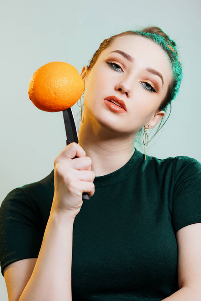 portrait of a beautiful young woman put a knife to an orange fruit ?? hands,fashion portrait on studio backgrond with green light - Fotó, kép