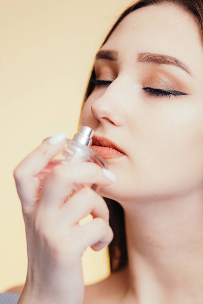 perfume bottle in the hands of a beautiful girl on a studio color background, part of the face of a young woman sniffing aroma, concept of female care and cosmetics - Photo, Image